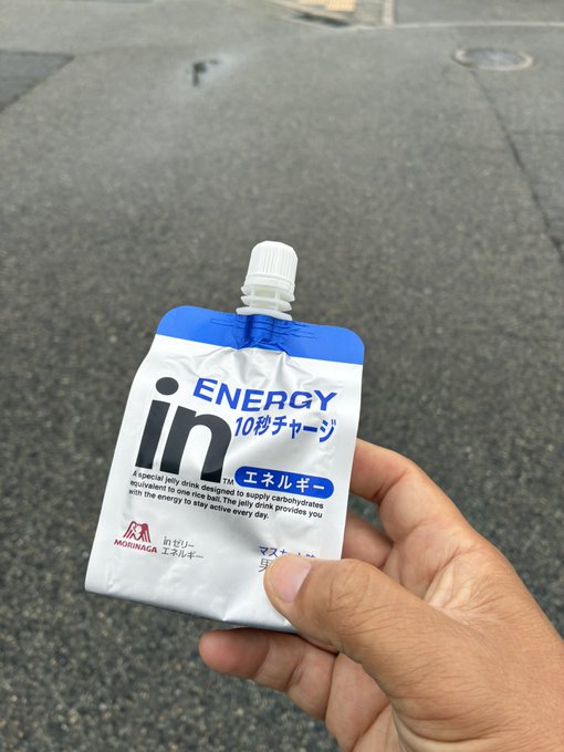 A hand holds up a pouch of "in ENERGY" jelly drink (10秒チャージ / エネルギー, by Morinaga) on a wet pavement after the rain has stopped, providing a quick carb boost before a 42 km #STEPN run.