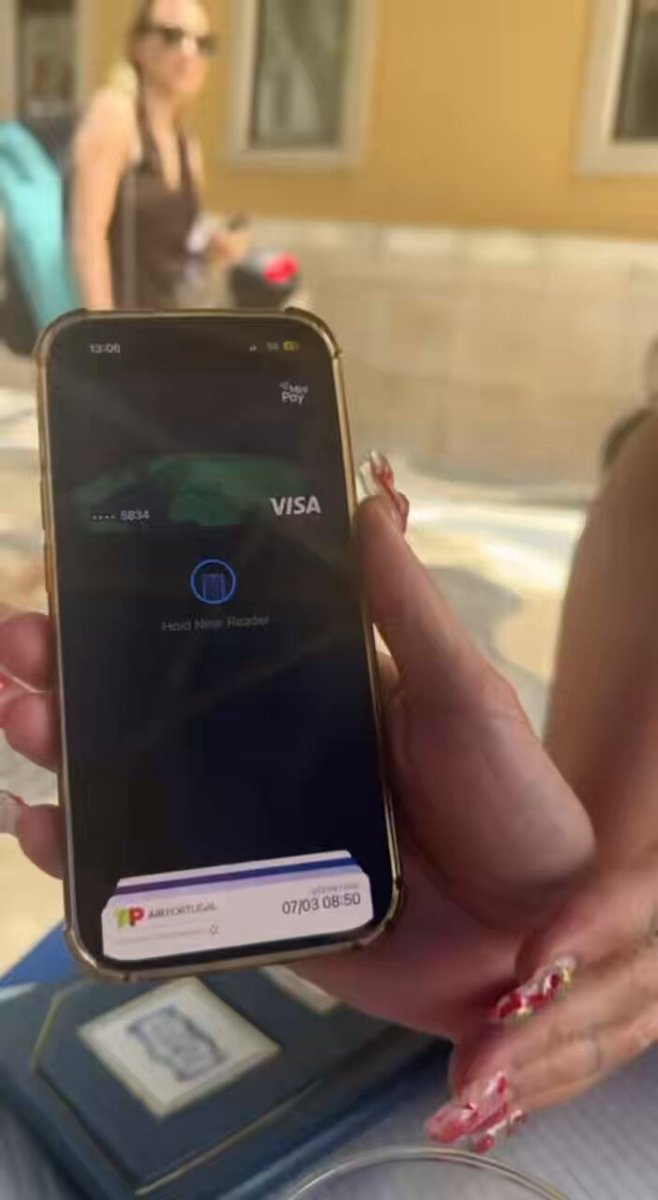 The image shows a person holding up an iPhone displaying a Visa card reader app. The person's hand is visible as they hold the phone with the app open. The background of the image features a building, suggesting that this scene takes place outdoors or in an urban setting.