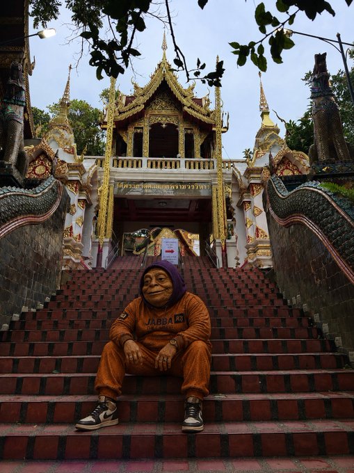 An older woman wearing a purple headscarf and orange sweatshirt sits on the red brick steps leading up to an ornate Thai temple with golden spires.