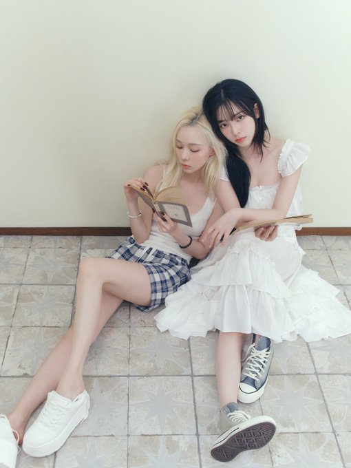 Two young women sitting against a plain wall on tiled floor, one with blonde hair wearing a plaid skirt and white tank top reading a book, the other with black hair in a white dress.