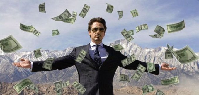 A man in a dark suit and sunglasses stands with arms outstretched as dollar bills rain down around him, set against a backdrop of snow-capped mountains and blue sky.