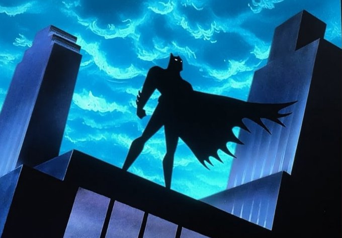 A stylized illustration of a silhouetted Batman figure with a flowing cape standing atop a building against a dramatic blue cloudy sky.