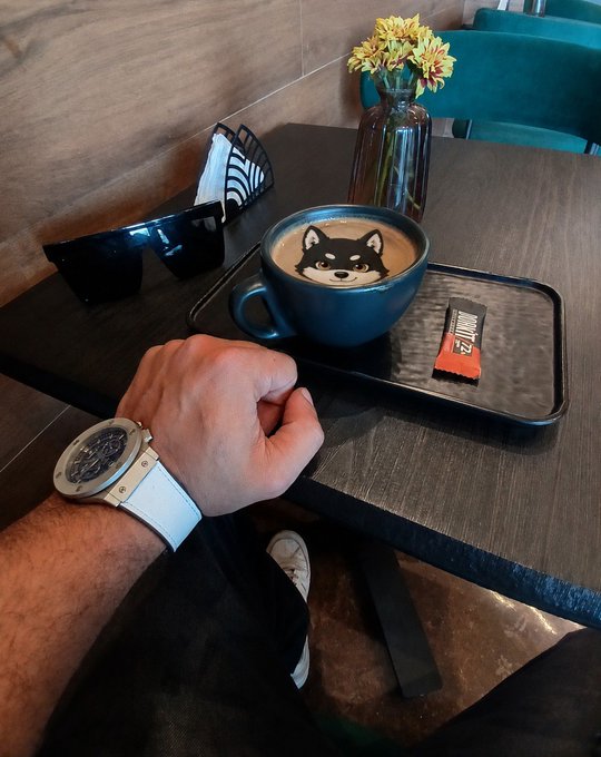 A first-person photo of a hand wearing a watch reaching toward a black coffee cup featuring Shiba Inu latte art on a dark tray.