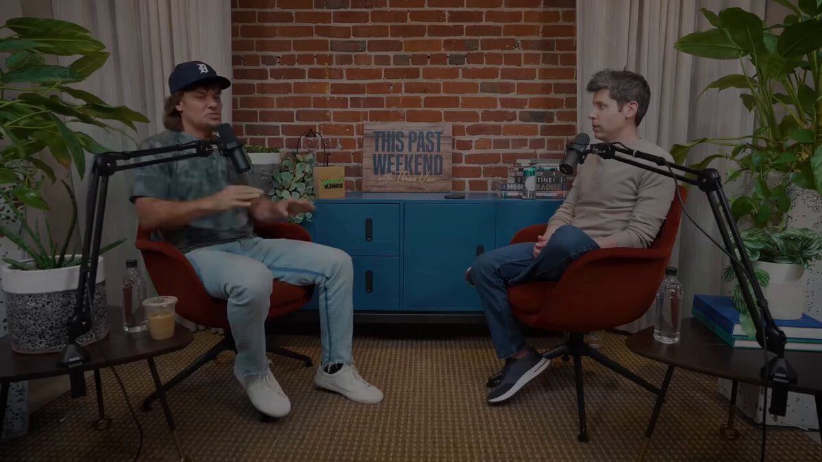 The image features two men sitting in chairs, engaged in a conversation. The man on the left is wearing a baseball cap and blue jeans, while the other man has short hair and is dressed more formally with a brown shirt and pants. They are both seated in front of a television set that displays an advertisement for 'Universal Basic Wealth'. This suggests that they might be discussing the content or implications of the ad, possibly related to wealth management or financial planning.

In addition to the chairs and TV, there is also a potted plant visible on one side of the image, adding some greenery to the scene. The presence of multiple books in the background further supports the idea that this could be an interview or discussion about personal finance or investing.
