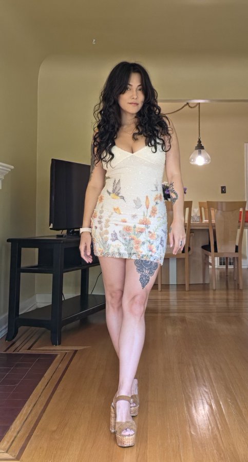 A woman with long curly black hair poses confidently in a cream-colored floral mini dress and tan platform heels in a living room setting. The caption "SOMEONE BUY ME A DRINKKK NOW $AVAGG" suggests a fun, flirtatious vibe, with her outfit and pose complementing the playful request for attention.