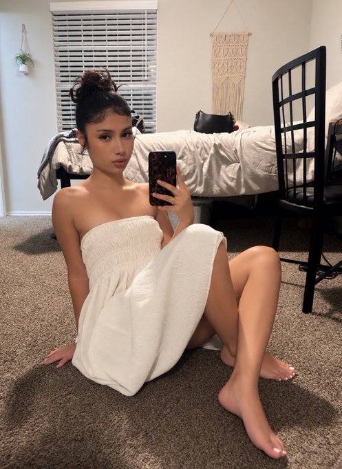 A young woman poses for a selfie in a white strapless dress, holding her phone with a leopard print case. The image, featuring a bedroom setting, complements the post's playful tone about the allure of certain aesthetics that can be both beautiful and expensive.