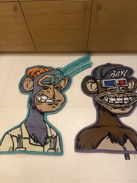 Two cartoon-style Bored Ape-style NFT rugs laid on a tile floor, one teal and one black, both with grinning expressions.