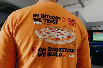 The image features a person wearing an orange shirt with a graphic design depicting a pizza. The design includes text related to Bitcoin and the concept of "on Bootstock We Build", which suggests that the person might be involved in the cryptocurrency or blockchain space, possibly as a developer or enthusiast.