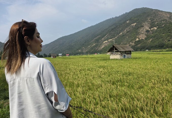 A woman with long brown hair wearing a white shirt stands with her back to the camera, looking out over a lush green rice field with a small wooden hut and forested mountains in the background.