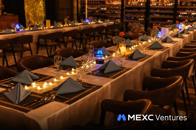 An elegantly set long banquet table featuring candles, folded napkins, glassware, and "MEXC" branded illuminated table tents, awaiting guests at the Alpha Arena 2026 Closing Gathering to celebrate the end of CoinfestAsia with an evening of networking and connections.