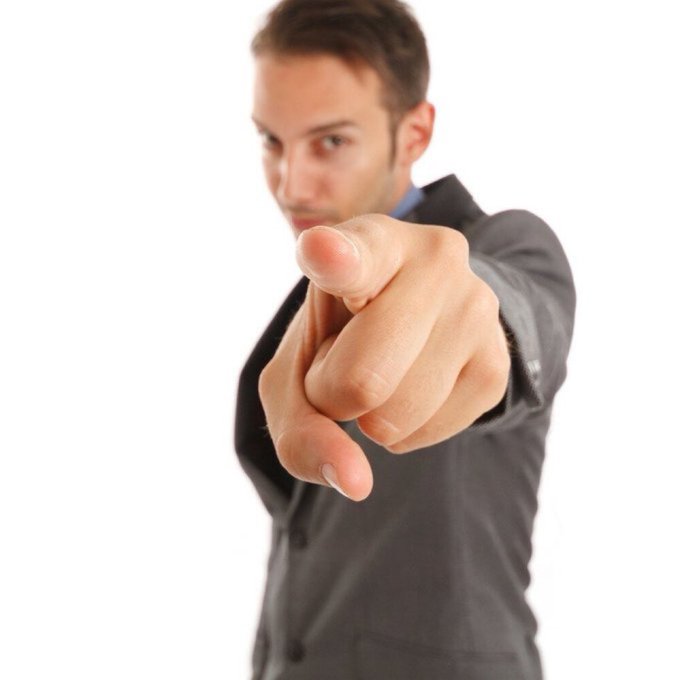 A man in a dark suit points directly at the camera with a serious expression, his finger in sharp focus against a white background.