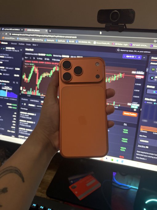 A hand proudly displays an orange iPhone 17 Pro Max in front of a computer monitor showing crypto trading charts (including Solana/SOL) on Kinesis, symbolizing a successful comeback from liquidation losses in DeFi liquidity providing with Meteora to finally afford the latest iPhone upgrade.