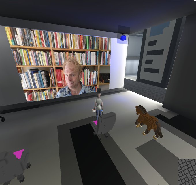 A virtual 3D environment showing a video projection of a man with a bookshelf background on a large screen, while small avatars and a brown animal stand in the room watching.