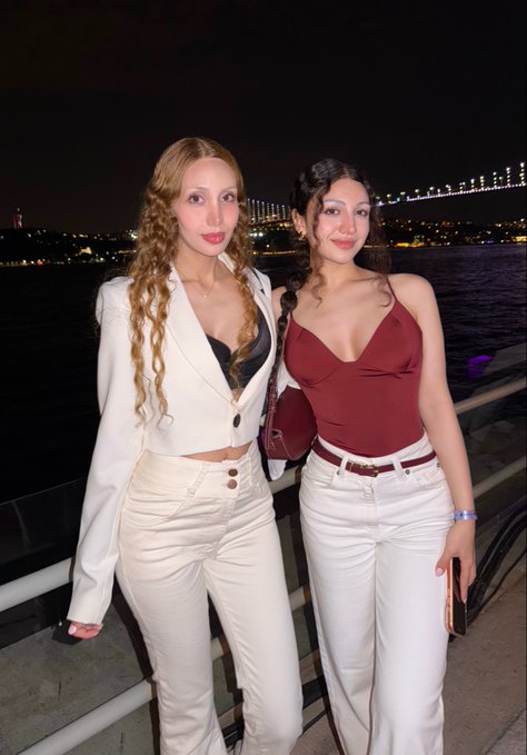 Two young women posing together at night near a body of water, with a lit suspension bridge visible in the background.