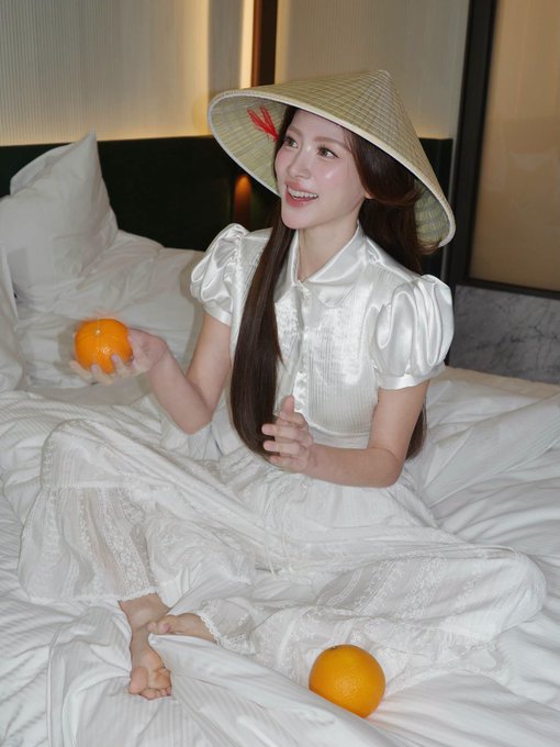 A young woman wearing a white dress and a traditional conical hat, smiling while sitting on a bed holding an orange.