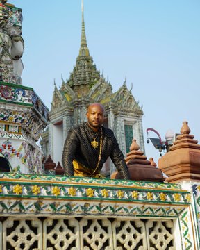 A man wearing a black patterned jacket and gold chains poses on an ornate, colorful structure at what appears to be the Grand Palace or a similar Thai temple complex with traditional spires in the background.