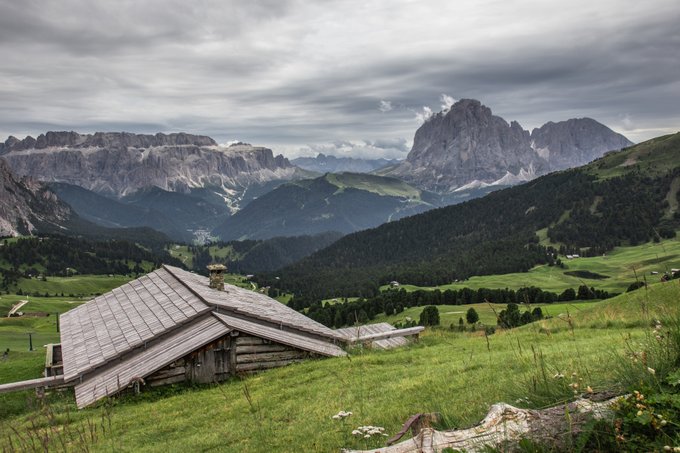 A scenic landscape photograph showing a rustic wooden alpine hut on a grassy meadow, with forested hills, dramatic mountain peaks, and a cloudy sky in the background.