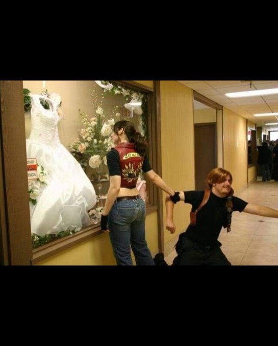 Two people are in a shopping mall corridor; one person in a cropped vest stands before a bridal shop window display, while another in dark clothing strikes a dramatic pose nearby.