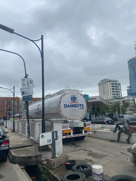 A tanker truck labeled "DANGOTE" is parked on a street under an overcast sky, with several tall buildings visible in the background and used tires stacked near the curb.