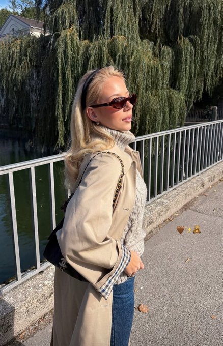 A blonde woman wearing sunglasses, a trench coat, and a white turtleneck stands on a bridge near a willow tree, with a small honey and bear emoji overlay.