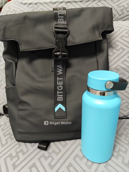 The image features a black backpack with a blue water bottle attached to it, sitting on top of a gray couch. The water bottle has a white label that says "bg钱包" and "bg u卡费率一样". The background is blurred, but the presence of the backpack and water bottle suggests they are placed in an indoor setting like a living room or office space.