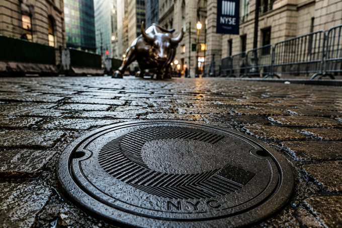 A low-angle shot focuses on a metal "NYC" manhole cover on a wet cobblestone street, with the bronze Charging Bull statue and an "NYSE" banner visible in the blurred background.