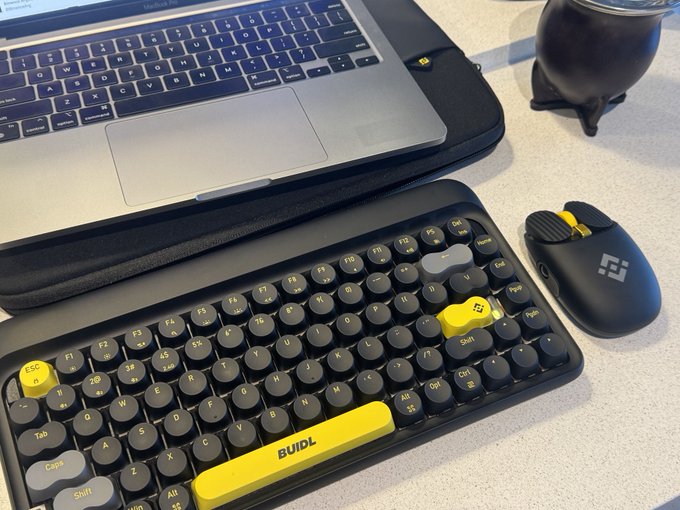 A desk setup featuring a laptop and a compact "BUIDL" mechanical keyboard with yellow accent keys, accompanied by an ergonomic mouse, captured as a "GMonday" morning greeting for the crypto community.