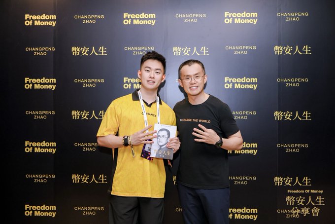 Two men pose in front of a step-and-repeat backdrop featuring the text "Freedom of Money," "CHANGPENG ZHAO," and the Chinese characters "幣安人生" (Binance Life), with the younger man on the left holding a copy of the book "幣安人生" featuring CZ's face on the cover. The image captures a fan meeting moment with Changpeng Zhao (CZ), the founder of Binance, as referenced in the post expressing gratitude for the conversation and humorously mentioning being blocked due to a malfunctioning insult robot.