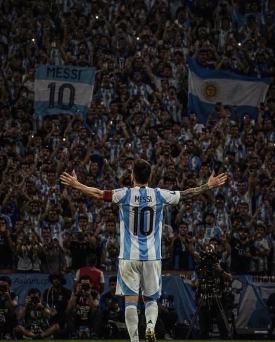 Lionel Messi wearing Argentina's number 10 jersey stands with arms outstretched facing a massive crowd, with an Argentine flag reading 'MESSI 10' and an Israeli flag visible among the fans.