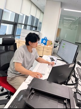 A candid photo of a young man in a grey t-shirt working at a desk with a laptop and monitor in a modern office setting, with a small robot mascot visible in the background.