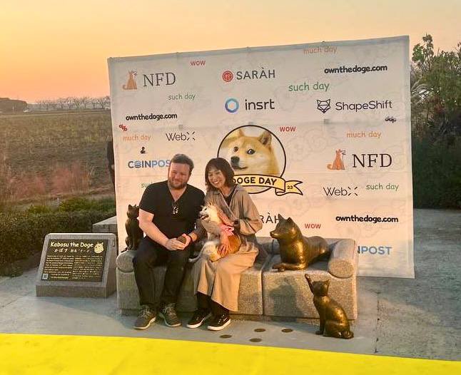 A couple poses with their dog "Kabosu the Doge" at the Doge Day "such day" event backdrop featuring sponsors like NFD, SARAH, insrt, ShapeShift, Web3, CoinPost, and ownthedoge.com, tying into the $OGDOGE tribute celebration. The sunset-lit scene commemorates the beloved Shiba Inu that inspired the Dogecoin mascot.