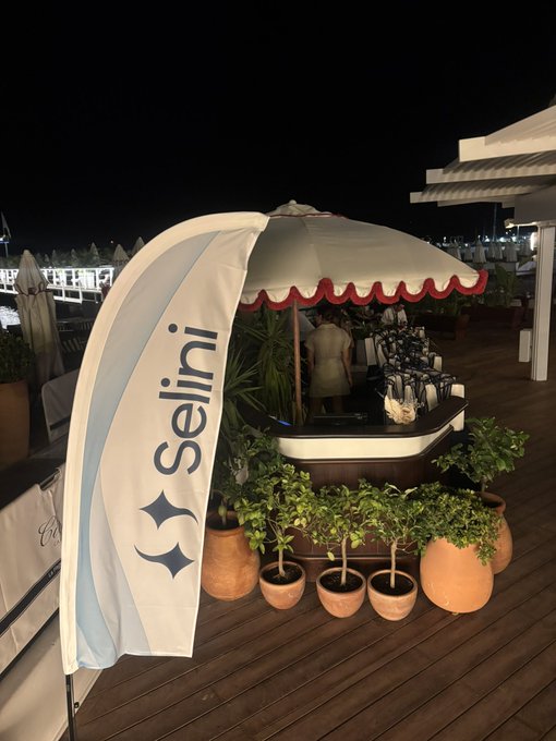 A nighttime outdoor scene shows a "Selini" branded banner flag next to an elegant cocktail bar setup under a scalloped canopy, with bottles and a bartender visible, reflecting the upscale investor dinner hosted by Selini Capital. Potted greenery surrounds the wooden deck venue, complementing the sophisticated atmosphere of the networking event.
