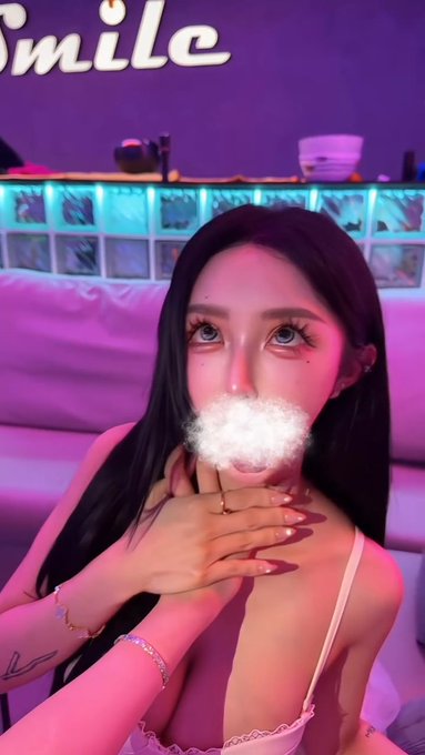 A young woman with long dark hair and blue eyes poses in a dimly lit, neon-colored room with the word 'Smile' displayed on the wall behind her, blowing a puff of vapor or smoke from her mouth.