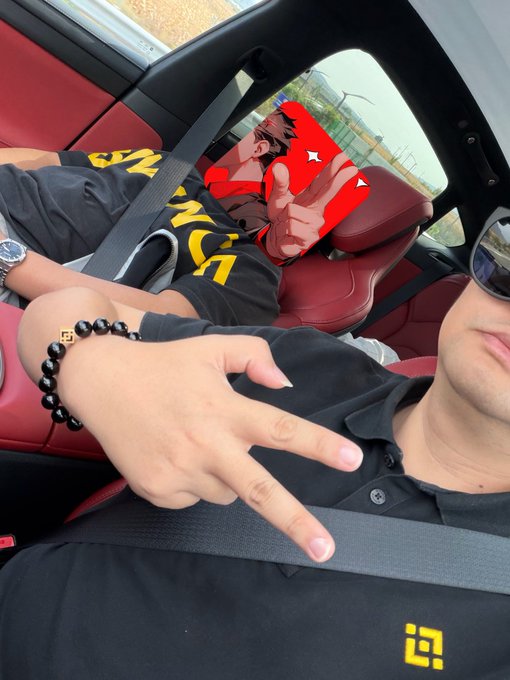 Two people sit inside a car with red leather seats; the person in the foreground wears a black polo shirt and makes a peace sign with their hand, while the other person's face is obscured by a digital overlay.
