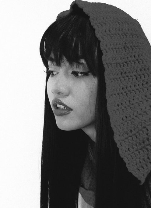 A black-and-white portrait of a young woman with long dark hair, striking eye makeup, and dark lipstick, wearing a textured knit hood against a white background.