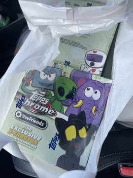 A white plastic shopping bag from "Veefriends" sits in a vehicle, containing items featuring cartoon characters including a green figure labeled "CHIP CHROMO" and a purple character.