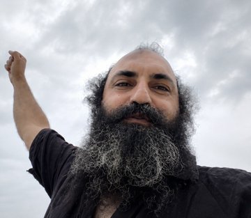 A low-angle selfie of a bearded man with curly hair smiling against a cloudy sky.