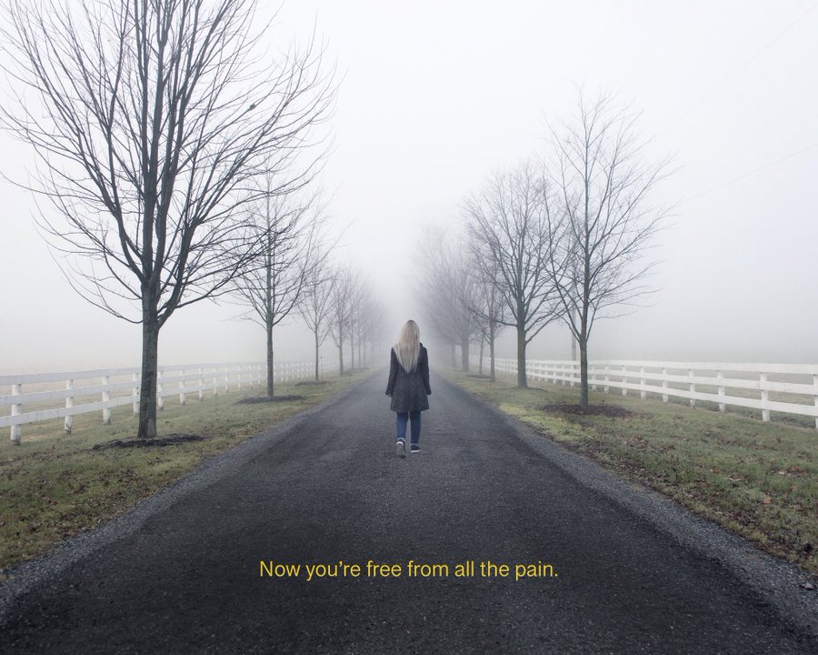 A blonde woman in a dark coat walks down a foggy, tree-lined road with white fences, accompanied by the text 'Now you're free from all the pain.'