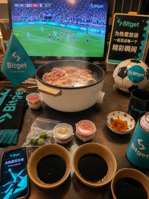 A promotional Bitget setup featuring a hot pot meal, a soccer ball, branded merchandise, and a TV showing an ARG vs FRA match.
