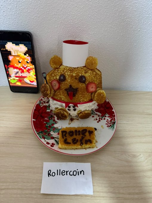 A teddy bear-shaped cake made for 'Rollercoin' sits on a decorative plate next to a smartphone displaying pixel art.