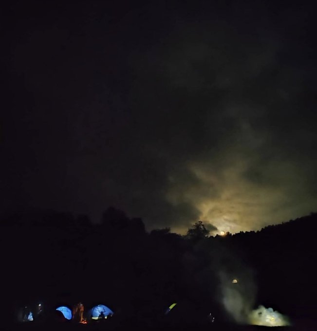 A nighttime scene of a campsite with illuminated tents nestled along a dark ridgeline, beneath a moonlit sky breaking through heavy clouds.