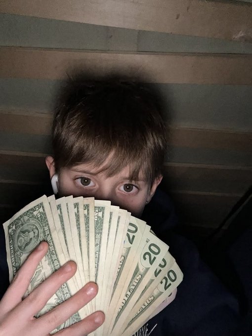 A young boy with brown hair holding a fan of US currency, prominently displaying multiple $20 bills and a $1 bill.