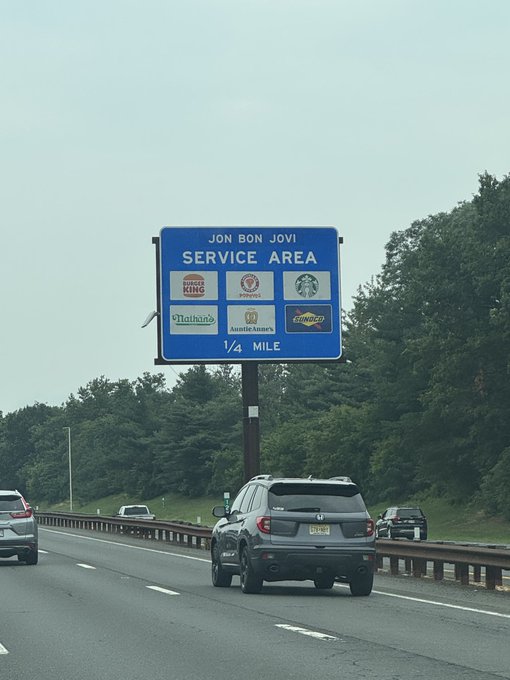 A highway sign for 'JON BON JOVI SERVICE AREA' 1/4 mile ahead, displaying logos for Burger King, Wendy's, Starbucks, Nathan's, Auntie Anne's, and Sunoco.