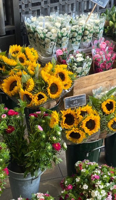 A display at a flower market featuring buckets of bright yellow sunflowers alongside bouquets of white, pink, and red flowers, with a small chalkboard sign reading 'Bouquets de 6 Bouquet'.