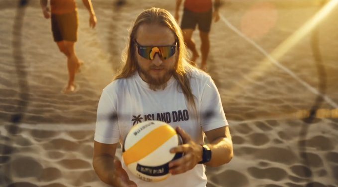 A bearded man with long hair wearing sunglasses and a 'JESUS AND DAO' t-shirt holds a volleyball on a sandy beach.