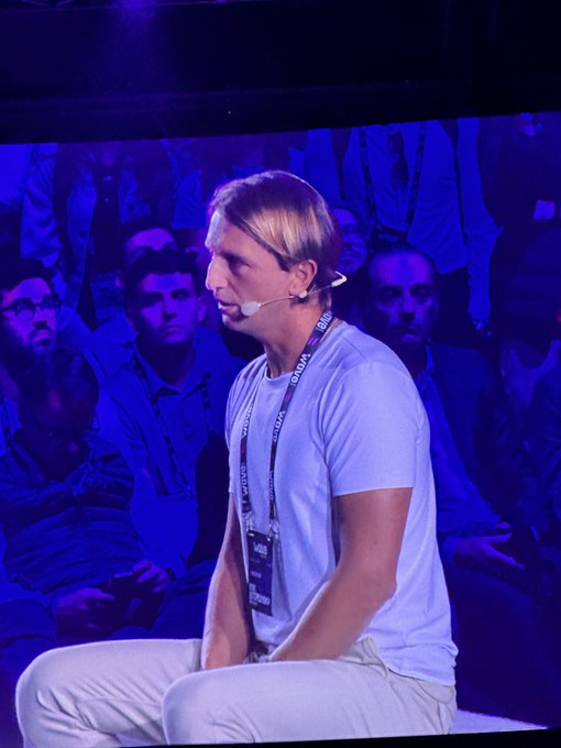 A photograph of a blonde man wearing a headset and a lanyard with 'RAFAEL' and 'AUTODESK' text, sitting cross-legged on a stage in front of an audience bathed in blue light.