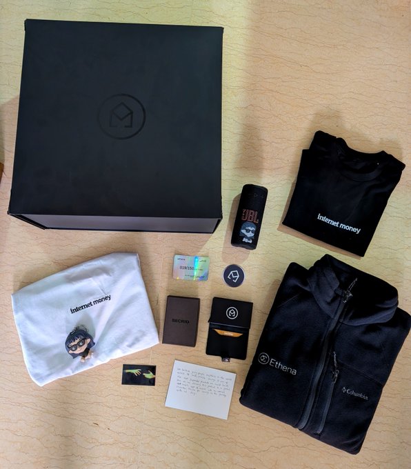 A flat lay photo shows an 'internet money' branded merchandise box containing a black t-shirt, white t-shirt with a cartoon character, JBL speaker, Etheos wallet, sticker, and black hoodie.