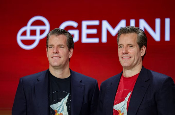 Two men are standing next to each other with their arms around one another, both wearing suits and smiling at the camera. They appear to be business partners or associates, as indicated by the name "Gemini" displayed above them on a large red sign. The image is related to a post discussing the partnership between GM and Gemini in relation to cryptocurrency, specifically Bitcoin futures. It seems that they are promoting the message of innocence or not being unfairly penalized for their actions with the previous accusation against them.