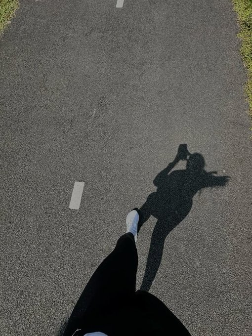 The image shows a person's shadow running down an asphalt road, with their feet positioned in front of them as they stride forward. The person appears to be wearing black pants and white socks while running. In the background, there are some grassy areas visible along the side of the road. The caption "Gm CT walking my own path at my own pace" suggests that this person is enjoying a leisurely run or walk in their preferred environment.