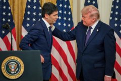 A photo showing two men in suits standing together with American flags in the background, with one man resting his hand on the other's arm.